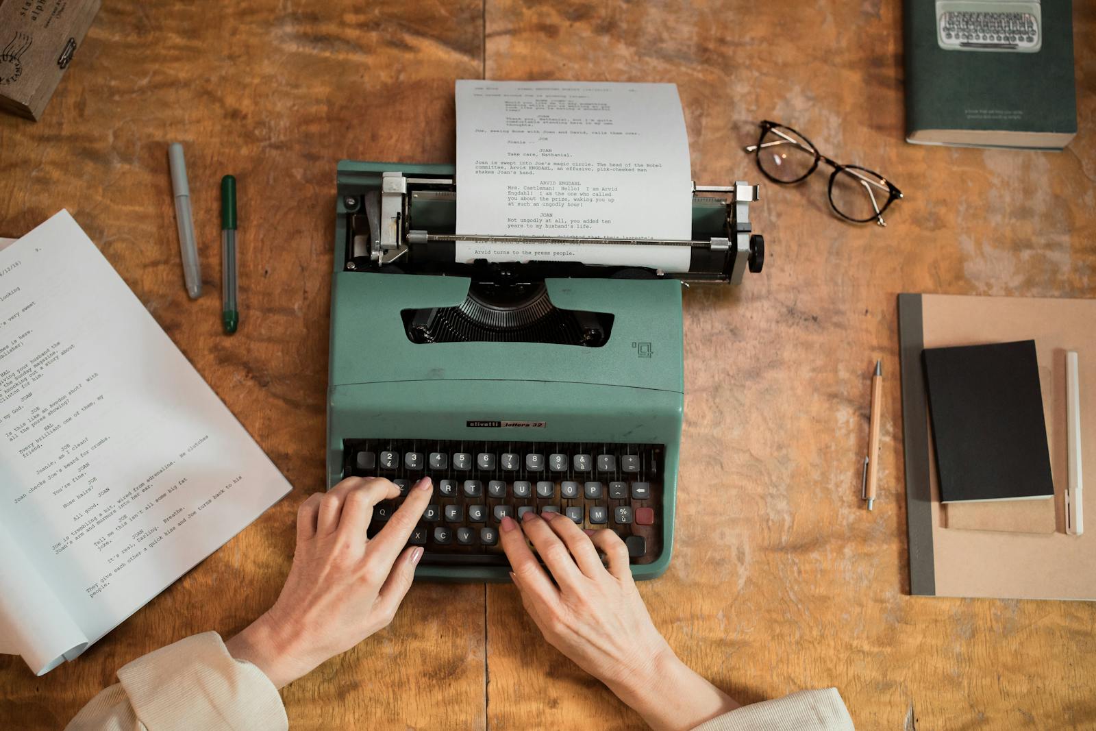 Hands working on a typewriter, paper drafts spread across a wooden desk