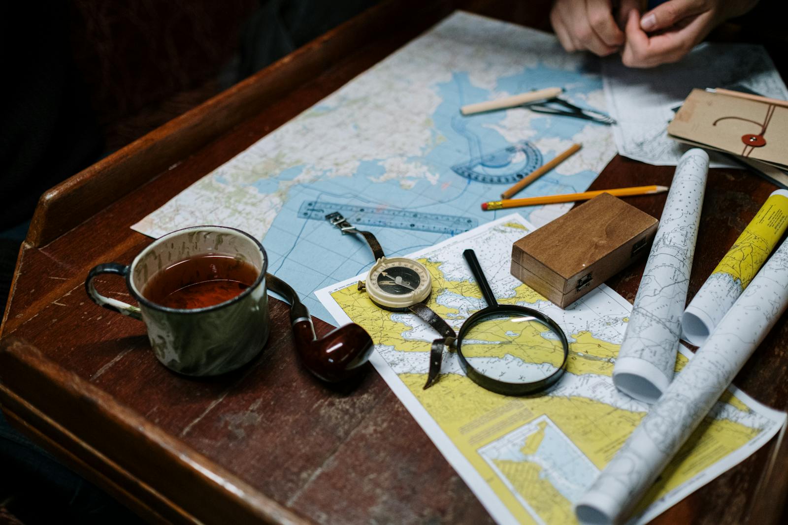 Maps and charts spread across a wooden desk with a brass compass, magnifying glass, and pencils
